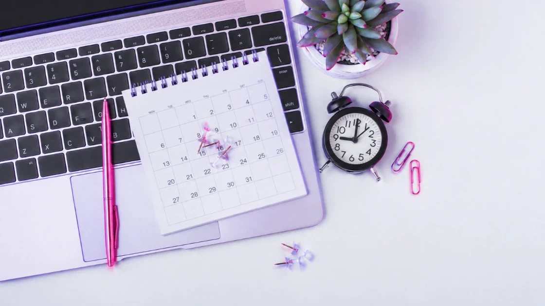 Bureaukalender met punaises op een laptop, naast een wekker en een roze pen.
